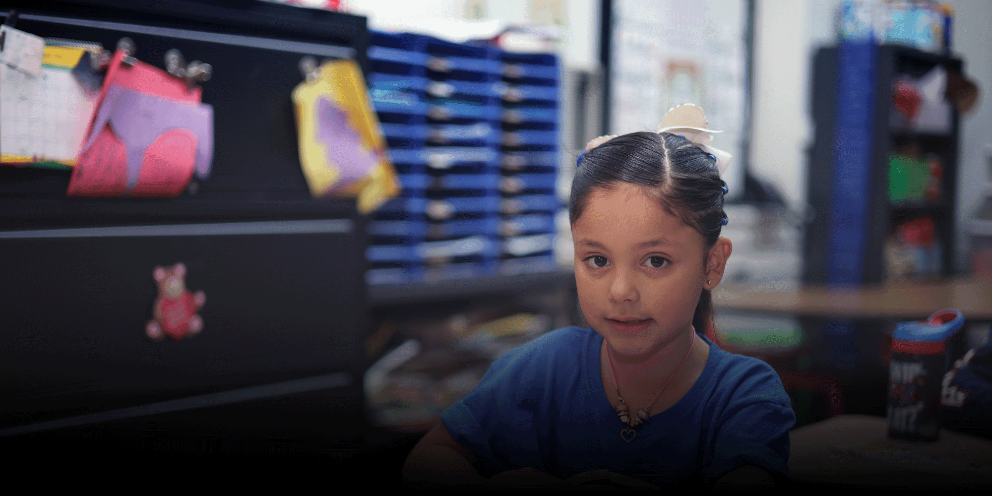 Student with a white hair bow seated at a classroom desk.
