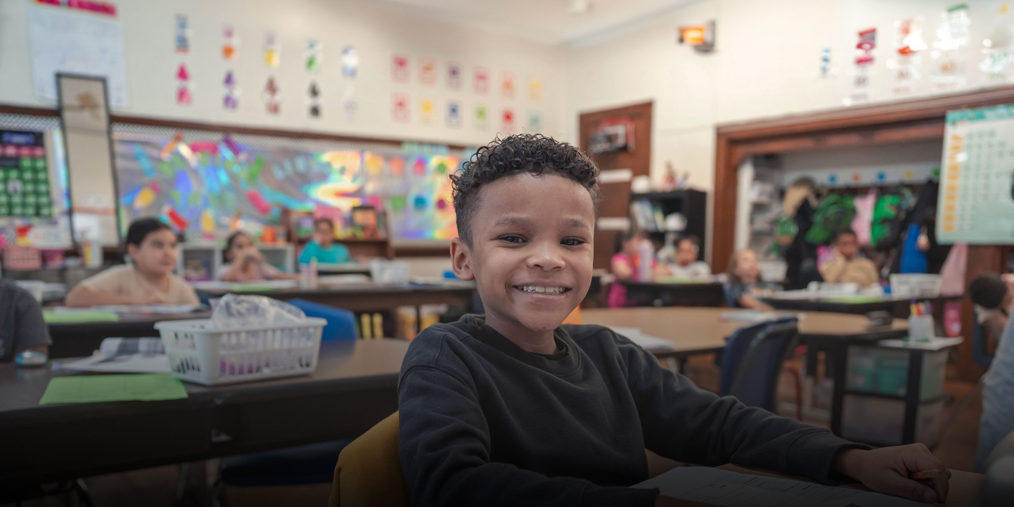 Smiling student seated in a classroom with classmates in the background.