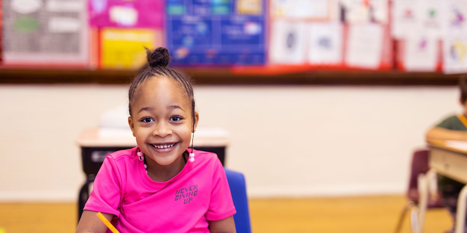 Smiling student writing with a pencil at their desk.